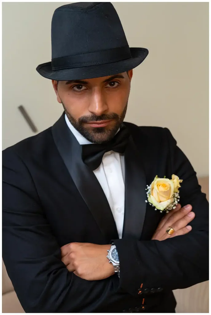 Der Bräutigam trägt einen schwarzen Hut und posiert mit verschränkten Armen im Hochzeitsanzug.
