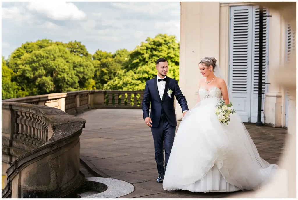 Brautpaar spaziert lächelnd über die Terrasse von Schloss Solitude, hält sich an den Händen und blickt sich verliebt an – die Braut im eleganten Hochzeitskleid, der Bräutigam im stilvollen Hochzeitsanzug.